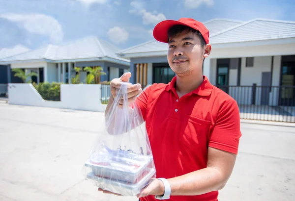 Asian delivery servicemen wearing a red uniform with a red cap and handling food boxes in plastic bags to give to the customer in front of the house. Online shopping and Express delivery