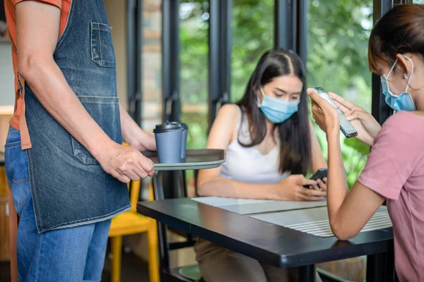 Asian woman with a friend playing on the phone and drinking hot coffee in a coffee shop being served drinks by the waitress and Both of you wear masks