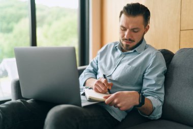 Sinirli esmer adam dizüstü bilgisayarla çalışıyor ve notlar alıyor.