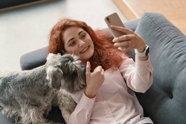 Kızıl saçlı ve sevimli gülüşlü bir kız kanepede uzanıyor ve evcil hayvanıyla selfie çekiyor.