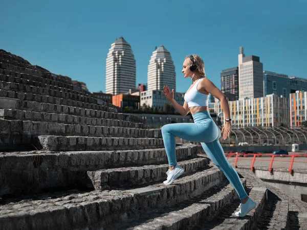 Beautiful fitness girl running on the city background in sexy blue leggings and earphones in the rays of the rising sun.