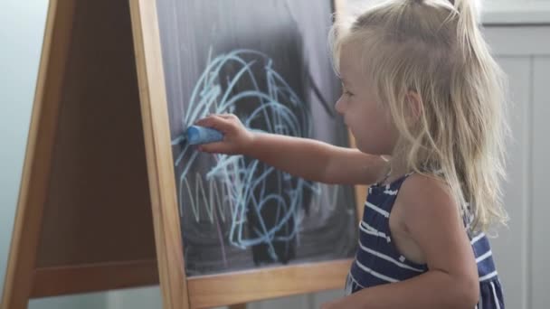 Den lilla flickan drar blå krita på en svart tavla. — Stockvideo