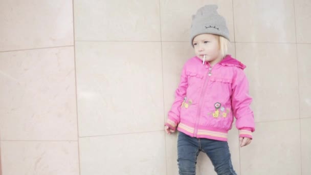 Een klein meisje is permanent op de muur van het huis en zuigen op een lolly snoep. — Stockvideo