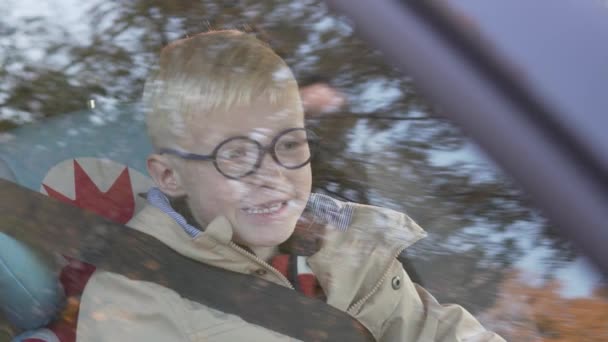 Een schooljongen bril zit in een auto in een autostoeltje — Stockvideo