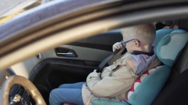 Gözlüklü bir çocuk arabanın ön koltuğunda bir araba koltuğuna oturur.