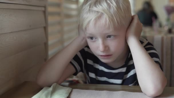Ein blonder Junge sitzt an einem leeren Tisch in einem Café — Stockvideo