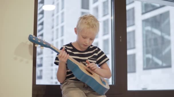 En ledsen pojke med en gitarr sitter vid fönstret — Stockvideo