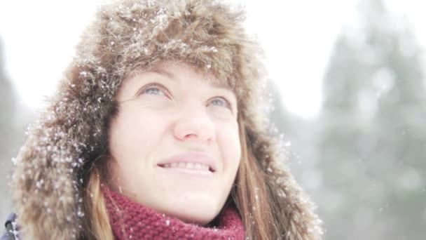 Uma jovem mulher em um chapéu de inverno quente com abas de ouvido olha para a neve caindo — Vídeo de Stock