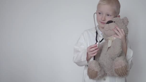 Rapaz bonito em um casaco de laboratório médicos ouvindo um ursinho de pelúcia com um estetoscópio — Vídeo de Stock
