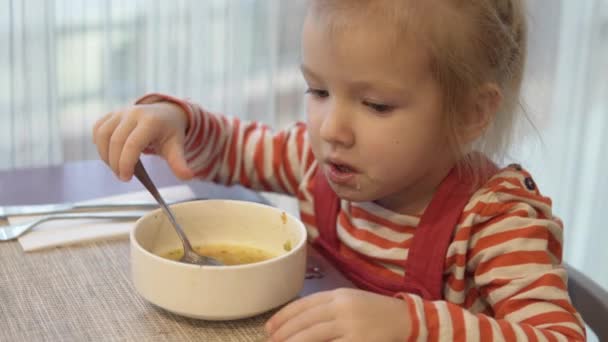 A kislány eszik zöldségleves. Ő ül az asztalnál, és tart egy kanál leves — Stock videók