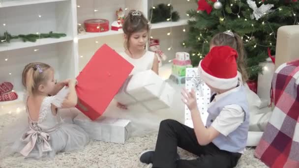 Элегантные дети сидят у елки и открывают красиво обернутые подарки — стоковое видео