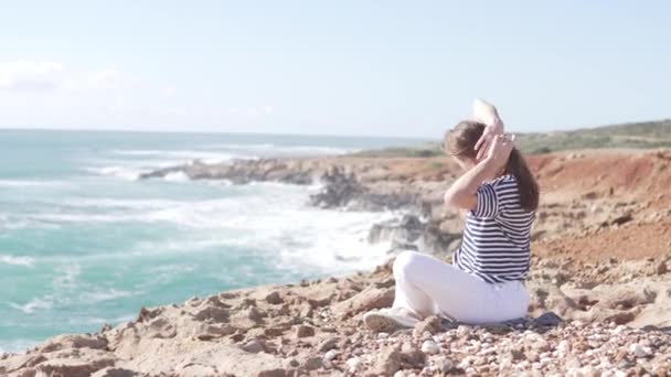 Молодая женщина сидит у моря и занимается гимнастикой — стоковое видео