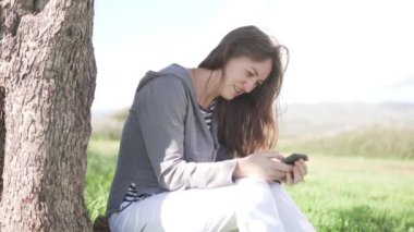 Genç bir kadın bir ağacın altında yeşil çim ente oturuyor ve telefona bakıyor.