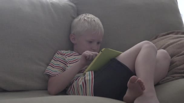O menino se senta no sofá e brinca no tablet — Vídeo de Stock