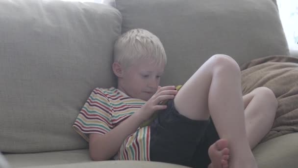 O menino se senta no sofá e brinca no tablet — Vídeo de Stock