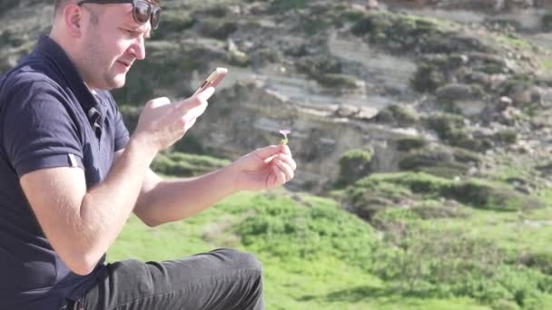 Чоловік фотографує квітку на телефоні на фоні моря — стокове відео