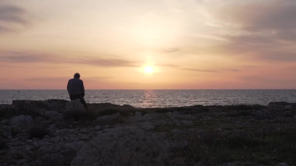Ein Mann sitzt und beobachtet einen Sonnenuntergang im Meer. — Stockvideo