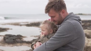 Küçük bir kız babasının kollarında oturur, denize bakarlar.