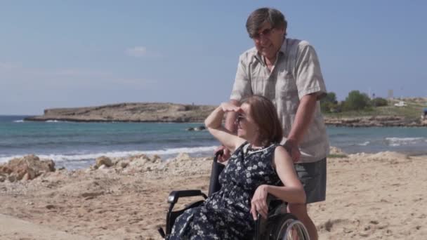 Een oudere man draagt zijn vrouw in een rolstoel langs de promenade — Stockvideo