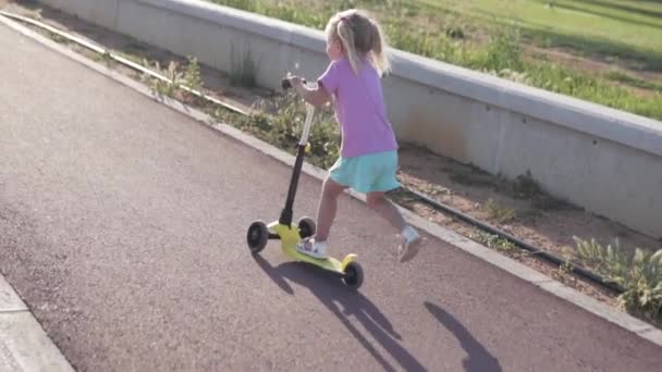 En liten flicka med två svansar rider en skoter i en Park — Stockvideo