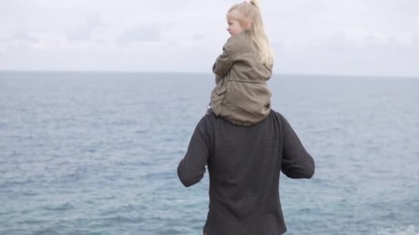 Маленькая девочка сидит на плечах своего отца и смотрит на море — стоковое видео