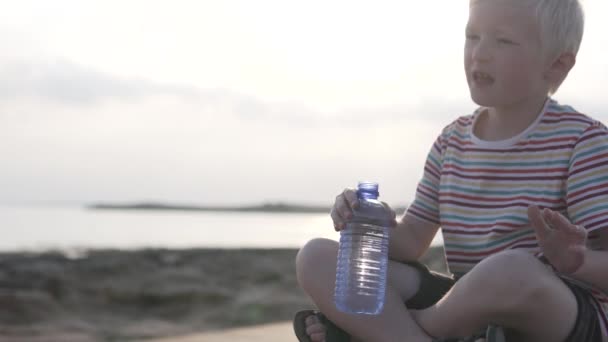 O menino bebe água ao pôr do sol junto ao mar . — Vídeo de Stock