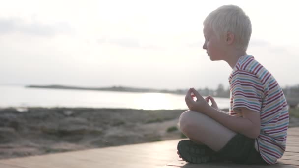 Un niño practica yoga en el fondo de una puesta de sol en el mar — Vídeos de Stock