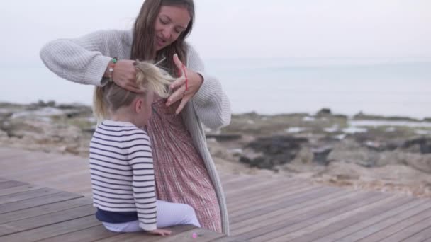 Mamma flätar håret på en liten flicka på stranden vid havet. — Stockvideo