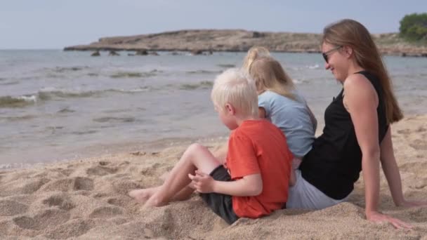 Schöne Mutter mit Kindern, die am Strand sitzt und aufs Meer blickt — Stockvideo