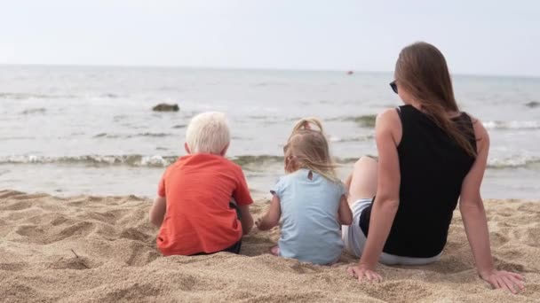 Vrouw met kinderen zit op het strand en kijkt naar de zee — Stockvideo