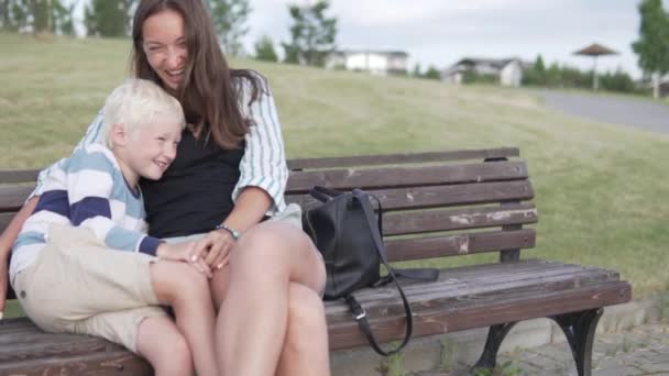 Mamá e hijo abrazan y ríen. familia se sienta en un banco en el parque de verano . — Vídeos de Stock