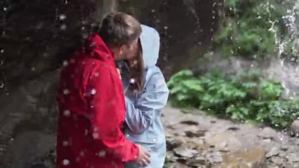 Um casal em uma caminhada fica sob um fluxo de uma cachoeira em jaquetas não molhadas , — Vídeo de Stock