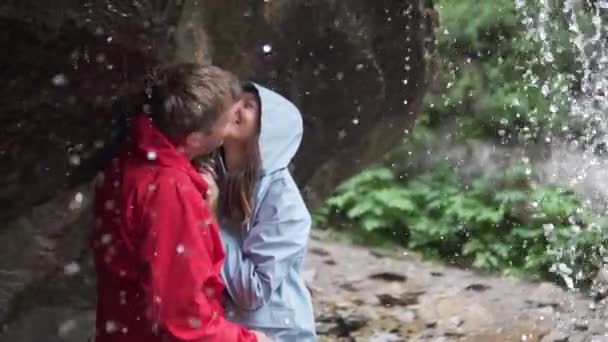 Casal em uma caminhada na montanha fica sob um fluxo de uma cachoeira em jaquetas não molhadas, eles gentilmente se beijam e riem — Vídeo de Stock