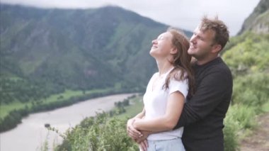 Bir adam bir kadına sarılır ve dağdan güzel bir manzaraya bakarlar.