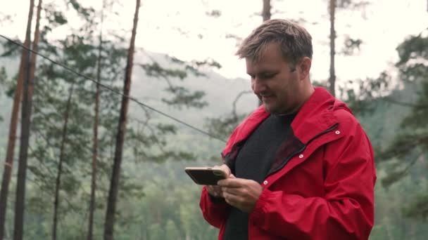 En man i en skog nära en fjäll älv tittar på en mobiltelefon — Stockvideo