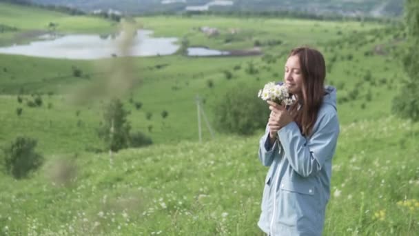 レインコートを着た若い女性が花を持って野原を歩く. — ストック動画