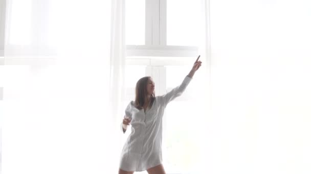 一个穿着睡衣的女人在早晨在窗边跳舞，窗子上长着白色的窗帘 — 图库视频影像