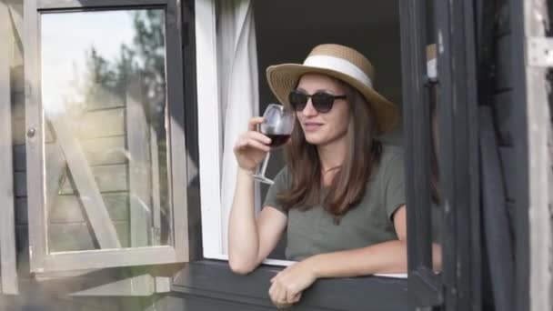 Una mujer con sombrero mira por la ventana con una copa de vino tinto — Vídeos de Stock