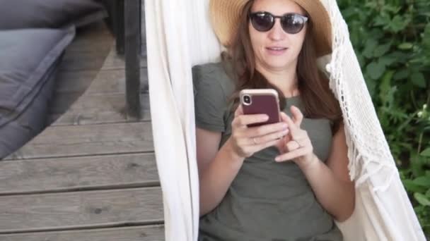 En kvinna vilar i en hängmatta och tittar på nyheter i en mobiltelefon — Stockvideo