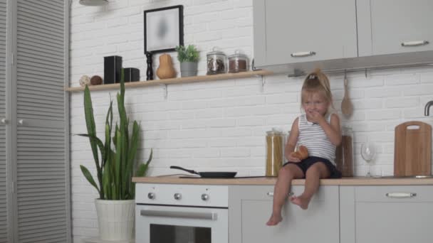 Funny little girl sitting in the kitchen and eagerly eating a loaf of white bread. — Stock Video