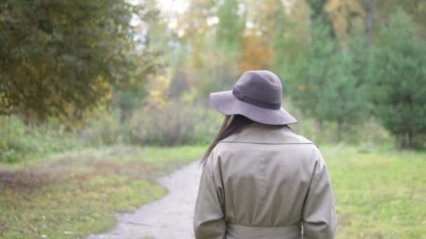 Жінка в капелюсі і плащі повільно йде через осінній парк — стокове відео