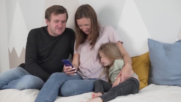 Familjen tittar på fotot på telefonen och tar en selfie hemma på sängen i barnrummet — Stockvideo