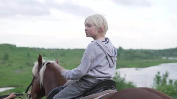 En vacker blond pojke rider en häst längs floden vid solnedgången. — Stockvideo