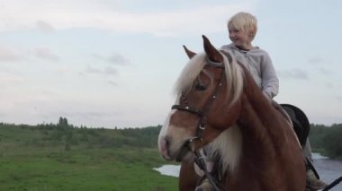 Yakışıklı sarışın bir çocuk gün batımında nehrin kenarındaki bir tarlada at sırtında oturur.