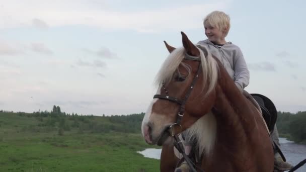 A handsome blond boy sits on horseback in a field by the river at sunset — Stock Video