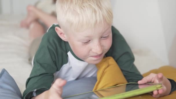 Um belo adolescente joga em um tablet em casa em uma cama — Vídeo de Stock