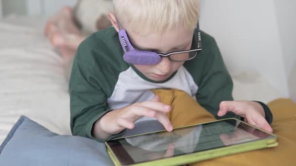 Um adolescente com óculos está envolvido em um tablet em casa na cama — Vídeo de Stock