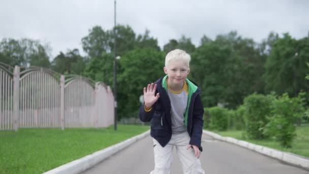 Belle adolescent garçon blond promenades sur patins à roulettes à la caméra et envoie un baiser d'air — Video