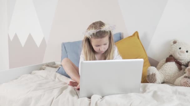 Graciosa niña escucha música en su computadora portátil en casa en la cama — Vídeo de stock