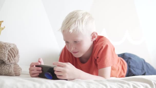 Lindo menino loiro joga o jogo em seu telefone celular em casa na cama — Vídeo de Stock